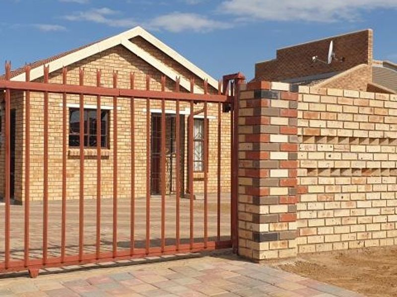 Front view of a face brick house featuring a secure metal gate, paved driveway, and a decorative brick boundary wall.