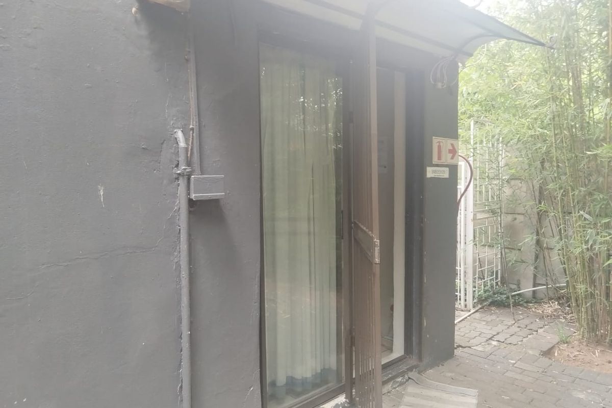 Exterior view of a guest house entrance featuring a security gate, glass sliding door, and paved walkway area with visible wear to the wall finish.