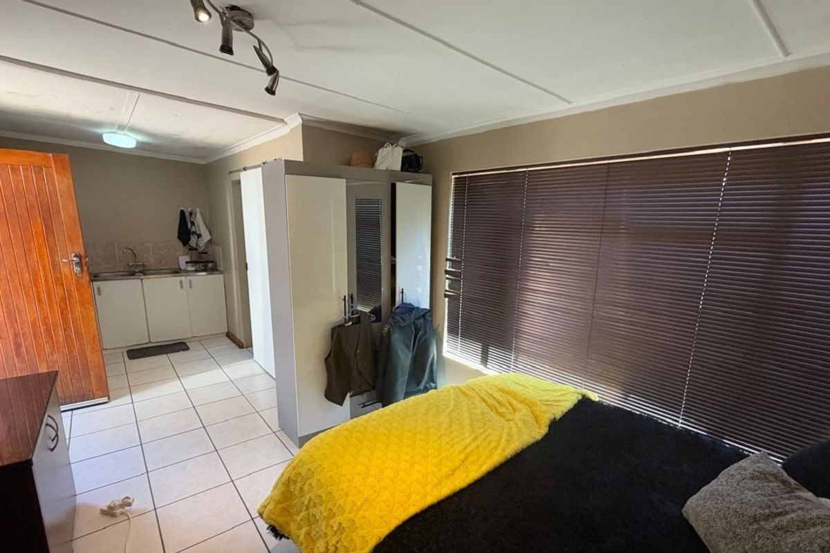 Open-plan studio apartment featuring tiled flooring, a compact kitchenette with built-in cabinetry, and large windows with venetian blinds.