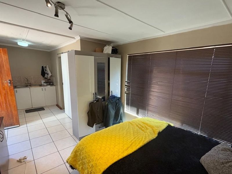 Open-plan studio apartment featuring tiled flooring, a compact kitchenette with built-in cabinetry, and large windows with venetian blinds.