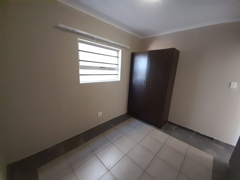 A simple, empty bedroom featuring tiled flooring, neutral walls, and a window fitted with security bars.