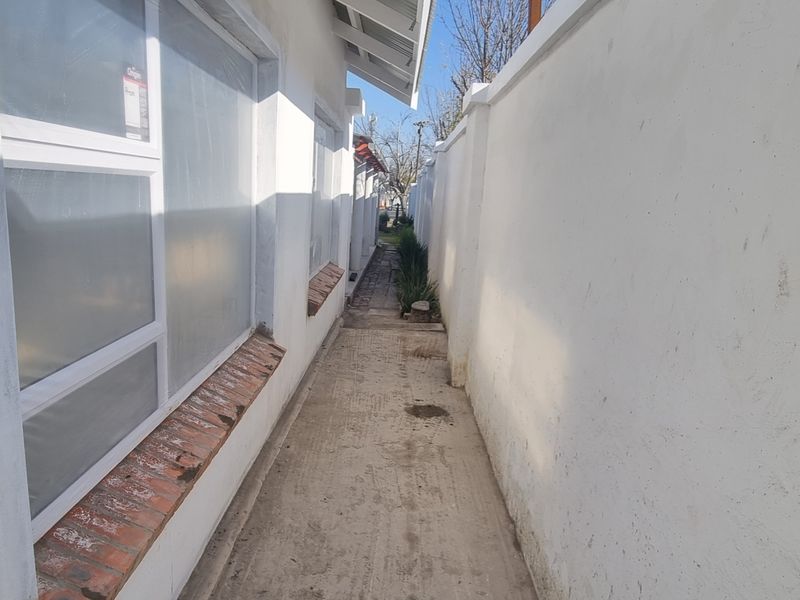A narrow side passage featuring a concrete pathway, boundary wall, and exterior windows with brick sills.