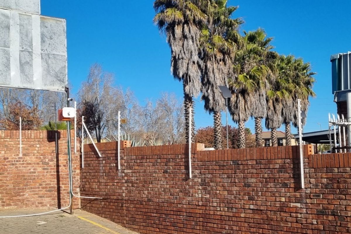Exterior view of a brick boundary wall featuring integrated electric fencing and security infrastructure, set against a backdrop of mature palm trees.
