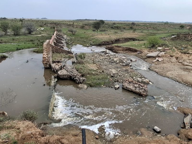 An elevated view of a farm landscape featuring a damaged stone weir structure across a natural watercourse with visible soil erosion.