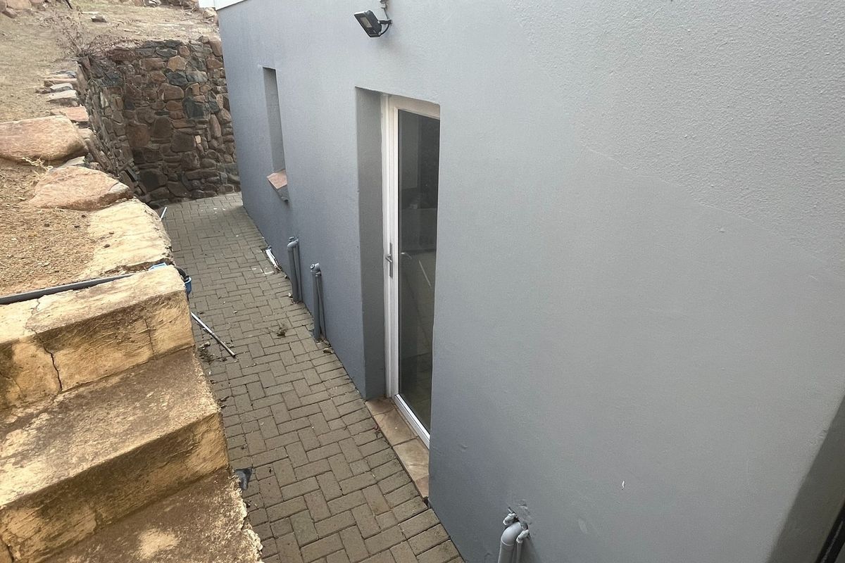 A side exterior view showing a paved walkway, a grey rendered wall, and a stone retaining wall structure.