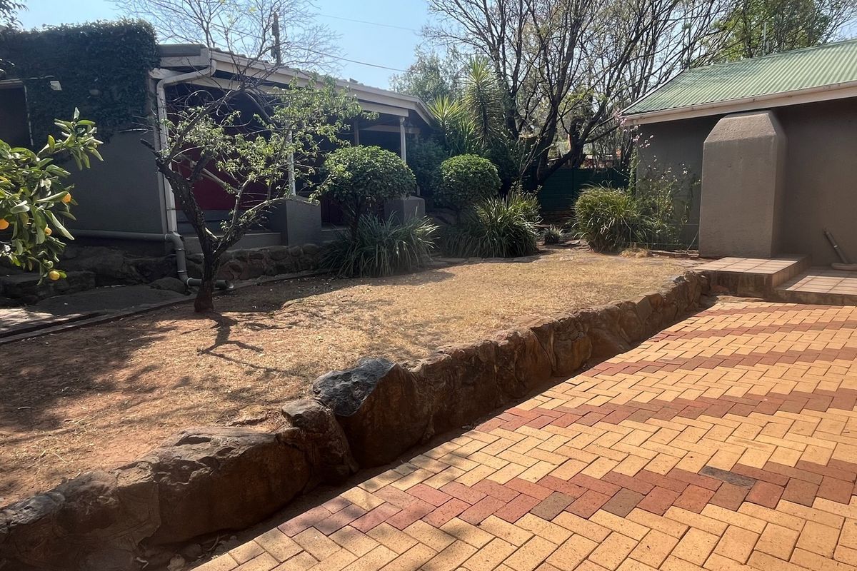 A rear garden area featuring a paved patio, stone retaining wall, and established landscaping between two building structures.