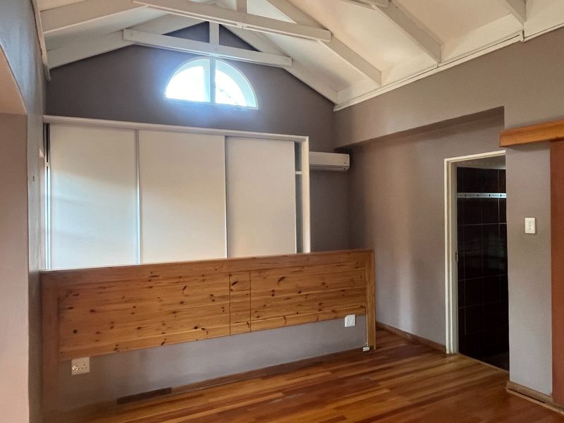 A spacious bedroom featuring vaulted ceilings with exposed beams, timber flooring, built-in cabinetry, and an arched window, leading to an en-suite bathroom.