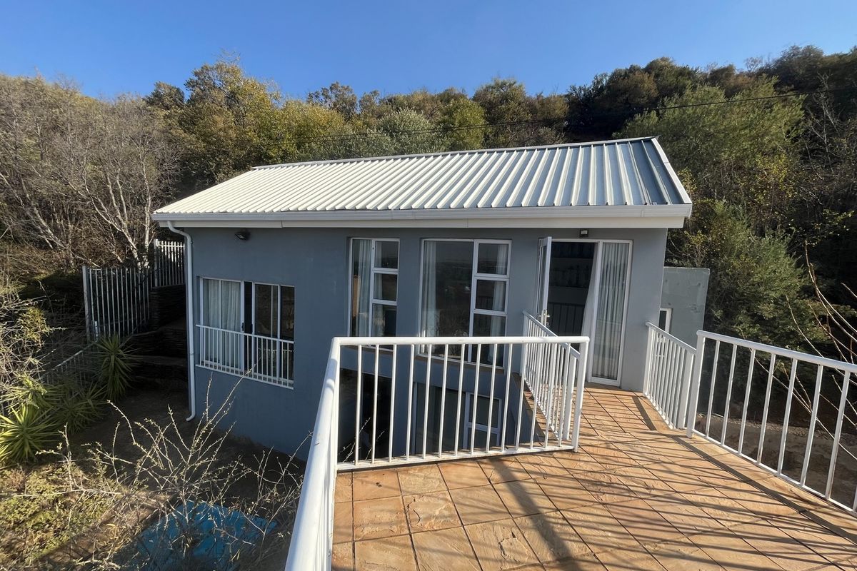 A detached exterior view of a single-story dwelling featuring a tiled balcony, metal roof, and white aluminum-framed windows set against a natural hillside.