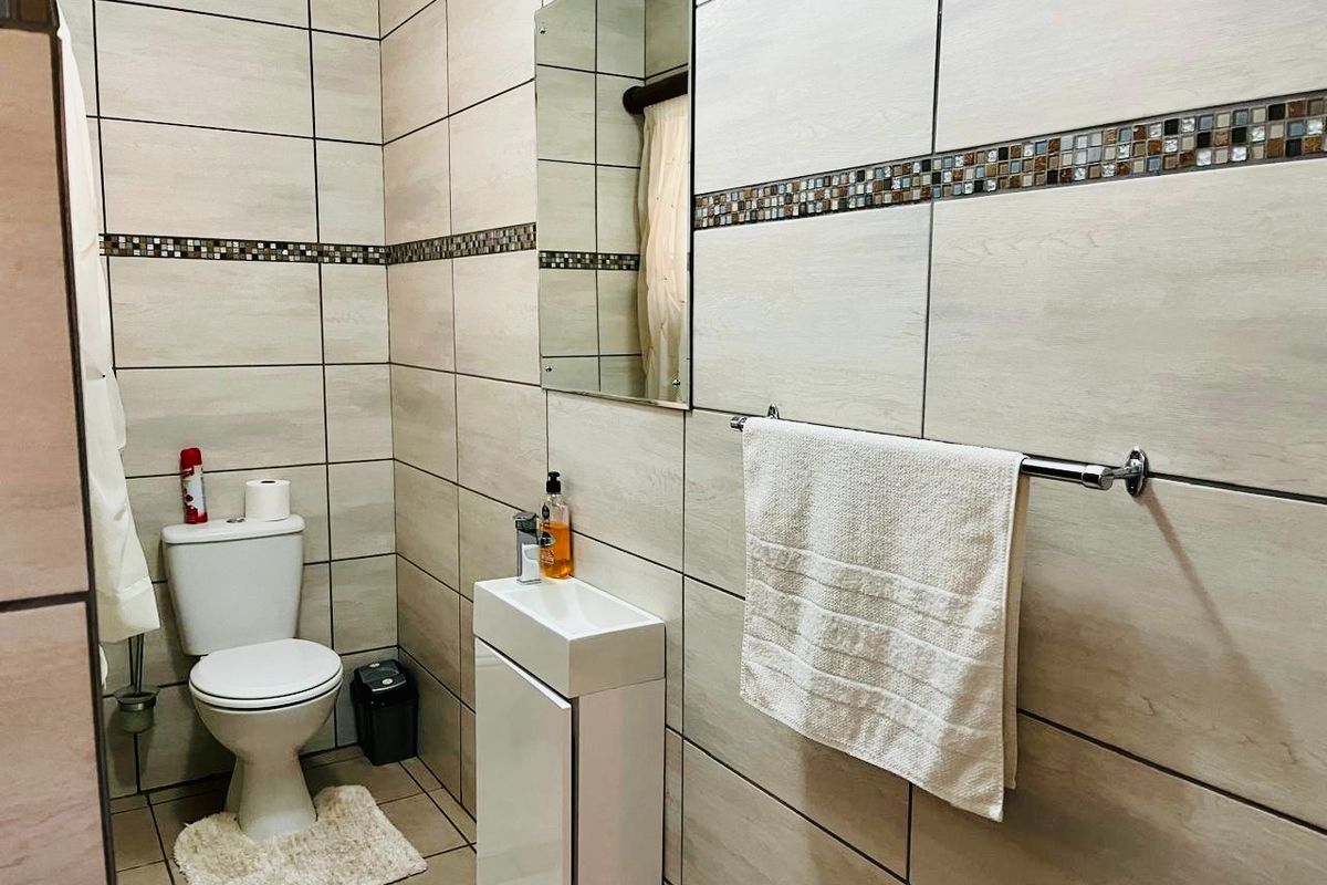 A compact, modern bathroom featuring neutral ceramic wall tiles with a decorative mosaic border, a wall-mounted basin with vanity, and a fitted mirror.