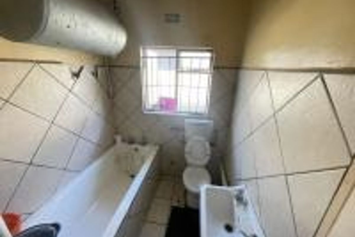 A functional bathroom featuring a bathtub, pedestal basin, and toilet, with tiled walls and an exposed electric geyser.