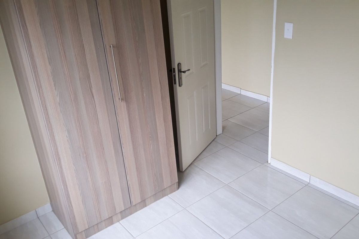 A bedroom featuring light-toned ceramic floor tiles and a built-in wooden wardrobe, leading into an adjacent hallway.