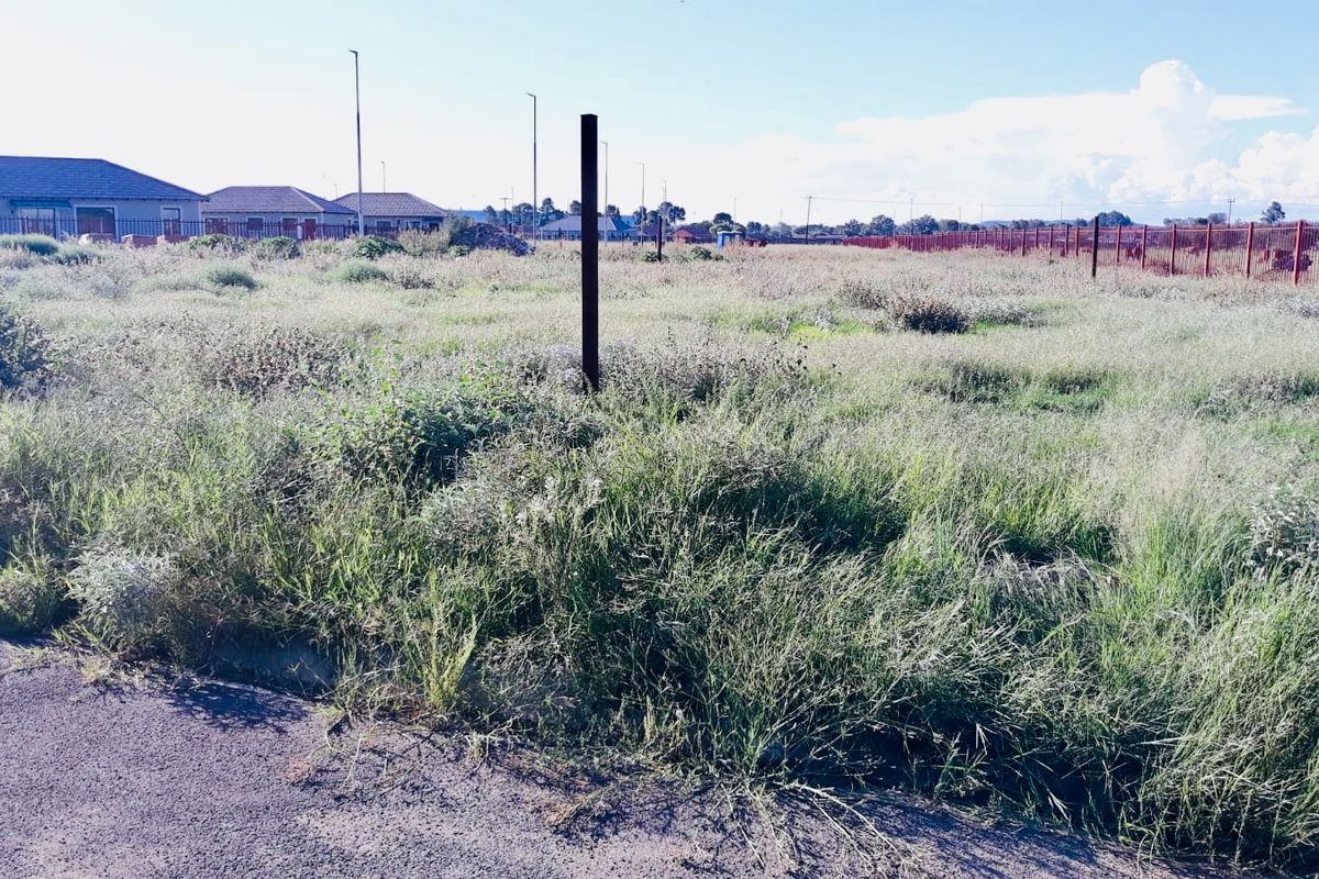 A vacant residential plot of land featuring overgrown grass, perimeter fencing, and direct street access in a developing neighborhood.