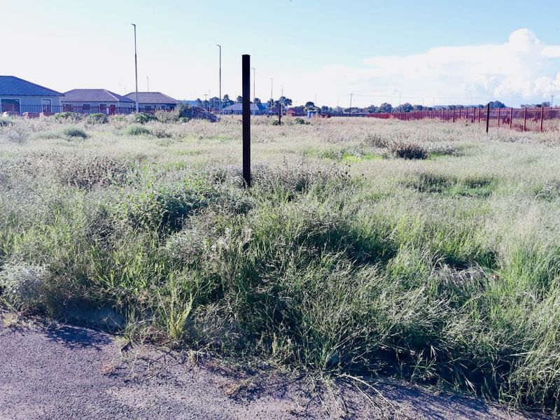 A vacant residential plot featuring overgrown grass and boundary fencing, situated with direct access to a paved road in a developed neighborhood.