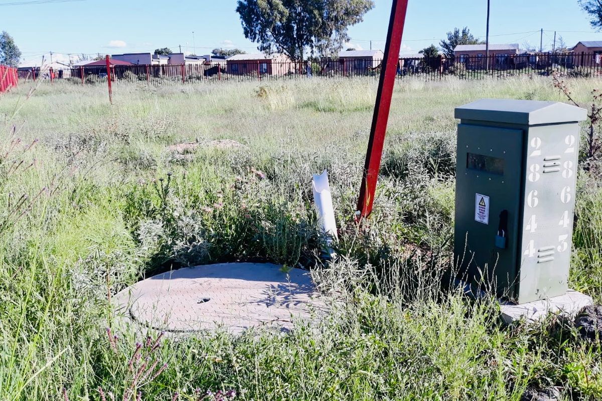 A vacant residential plot featuring an electrical distribution box, a manhole cover, and perimeter fencing, surrounded by overgrown grass.