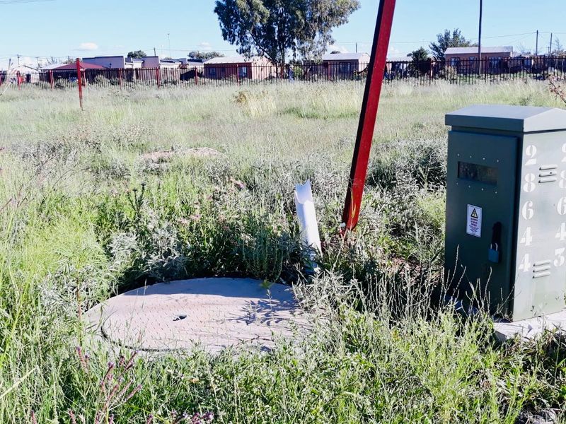 A vacant residential plot featuring an electrical distribution box, a manhole cover, and perimeter fencing, surrounded by overgrown grass.