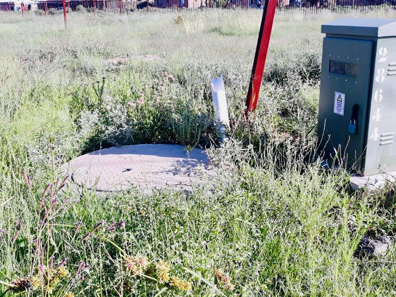 A vacant residential plot featuring an electrical distribution box and a manhole cover set within overgrown grass.