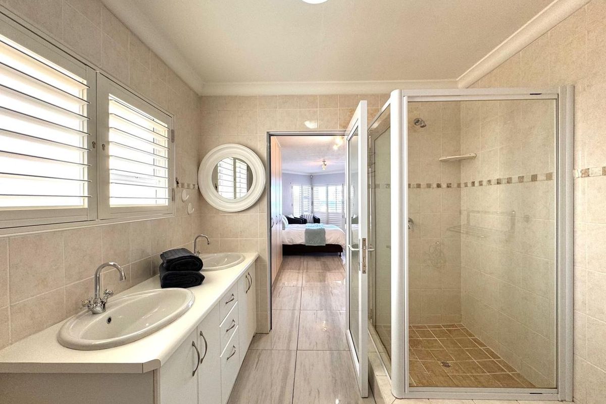 A well-maintained bathroom featuring a double vanity with integrated sinks, a glass-enclosed shower, and neutral floor-to-ceiling tiling with shutter blinds.