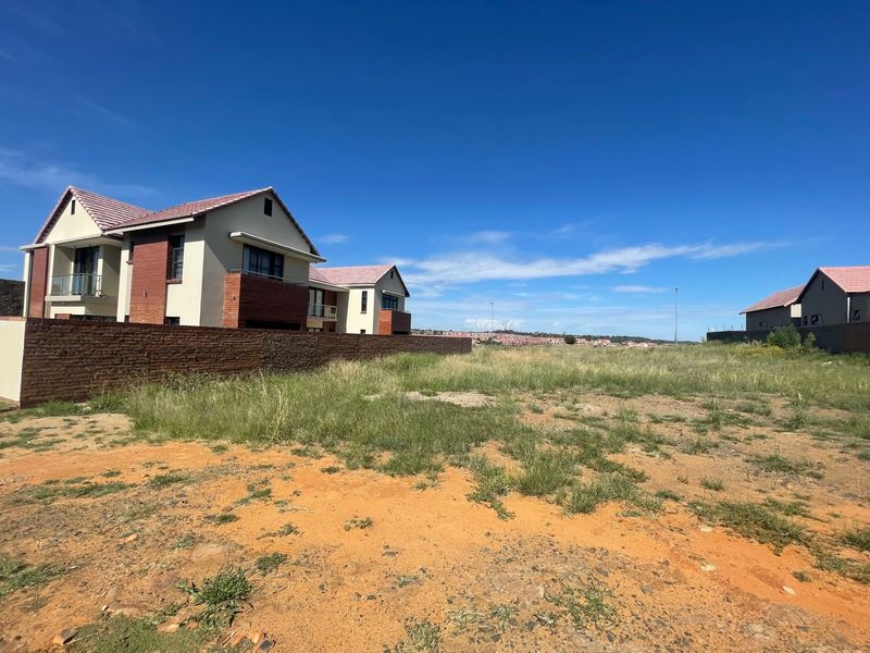 A vacant residential plot featuring natural grass and soil, adjacent to existing multi-story residential buildings with brick and plaster finishes.