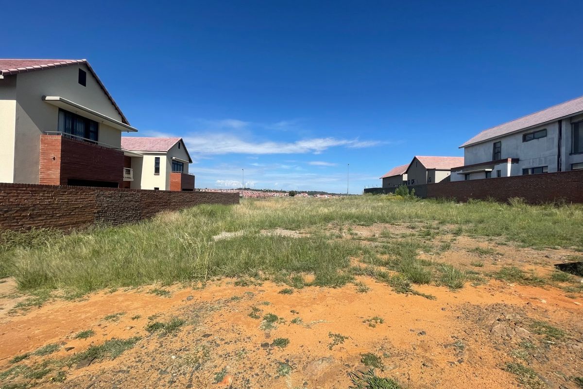 A vacant residential plot situated between existing modern homes, featuring boundary walls and natural ground cover.