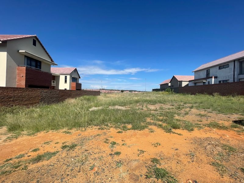 A vacant residential plot situated between existing modern homes, featuring boundary walls and natural ground cover.
