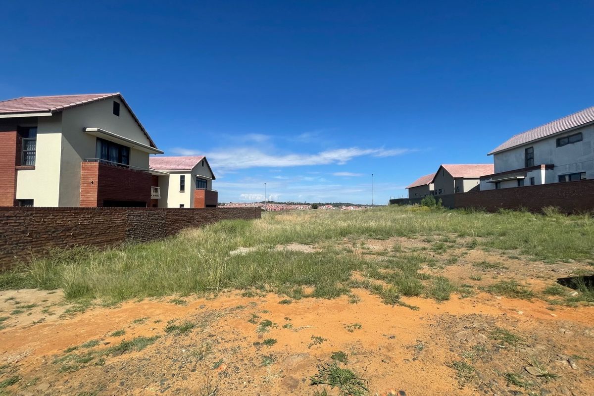 A vacant residential plot situated between existing modern homes, featuring a brick boundary wall and natural grass terrain.