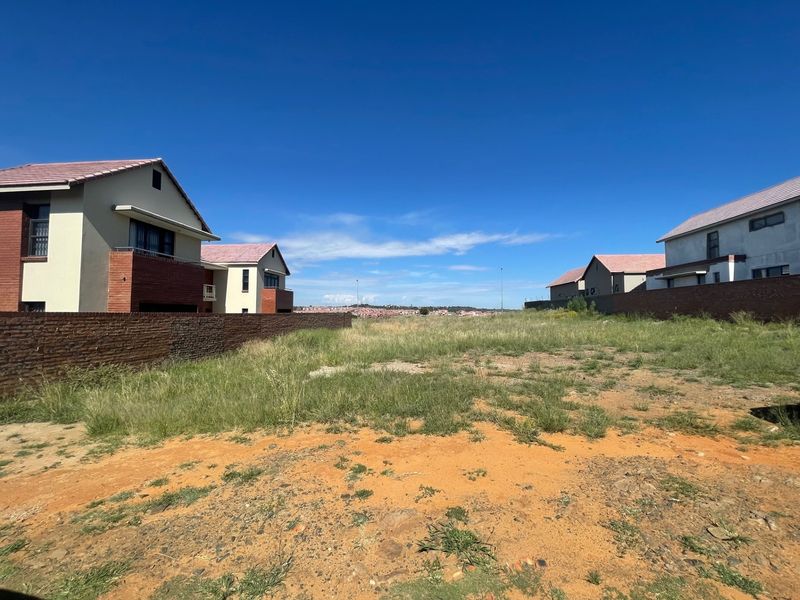 A vacant residential plot situated between existing modern homes, featuring a brick boundary wall and natural grass terrain.