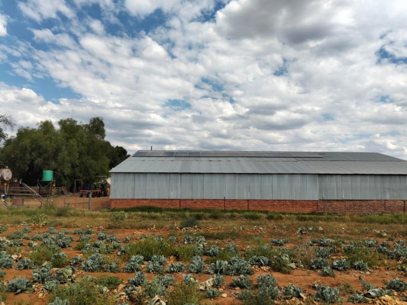 A view of a farm outbuilding featuring a corrugated metal roof with installed solar panels and a brick foundation, set against a cultivated field.