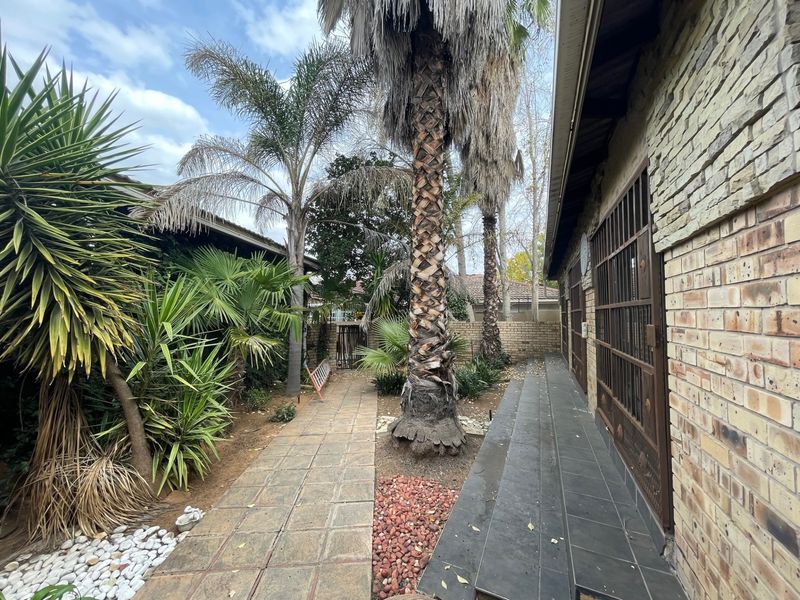 A rear exterior view featuring a brick and stone facade, a paved walkway, and established palm trees along the side of the property.