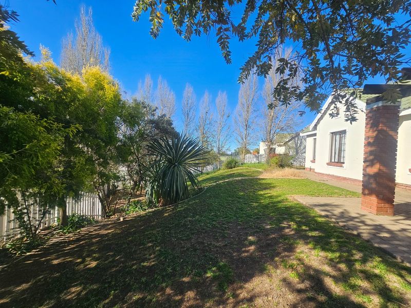 A rear exterior view of the townhouse featuring a paved patio area, brick pillar, and a spacious garden enclosed by a white picket fence.