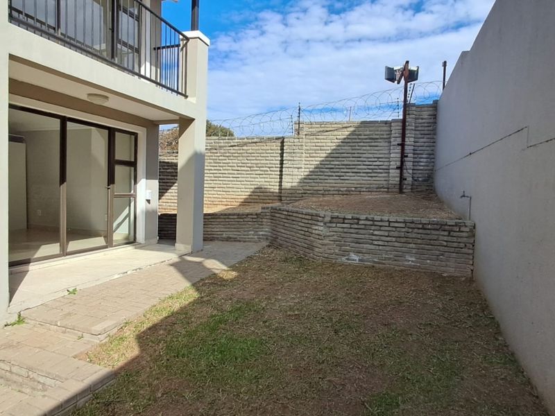 Rear exterior view of a townhouse featuring a paved patio, small garden area, retaining walls with security fencing, and an upper-level balcony.