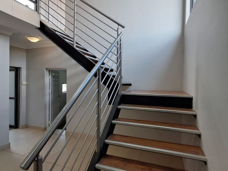 A bright, modern hallway featuring a staircase with wooden treads and a sleek stainless steel balustrade, leading to an open landing area.