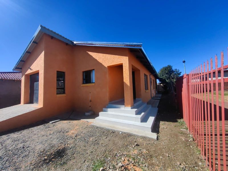 Front view of a single-story house featuring textured orange plaster walls, tiled entrance steps, and a secure metal perimeter fence.