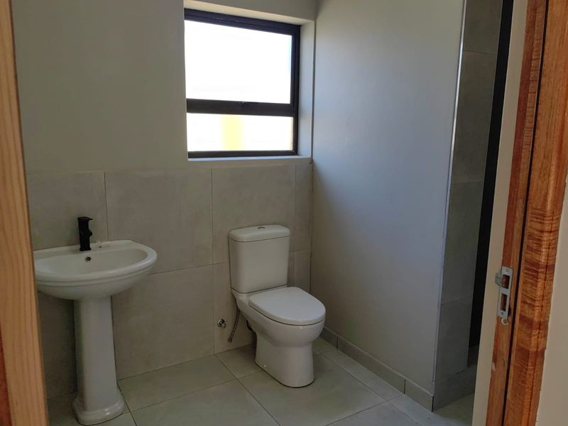 A clean, modern bathroom featuring a pedestal basin, toilet, and neutral wall and floor tiling with a window for natural light.