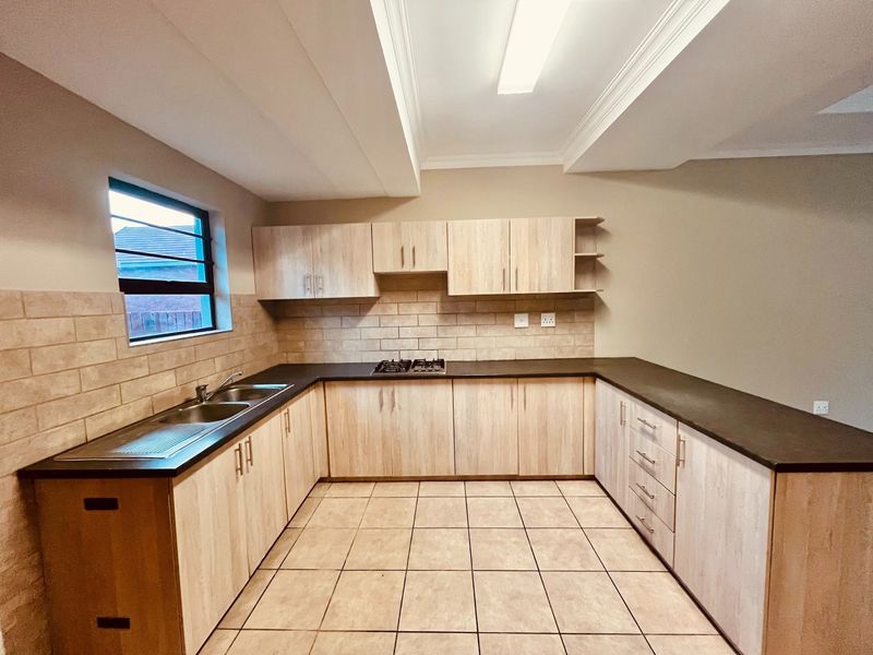 A clean, functional kitchen featuring light wood-finish cabinetry, dark countertops, a gas hob, and neutral tiled flooring and backsplash.