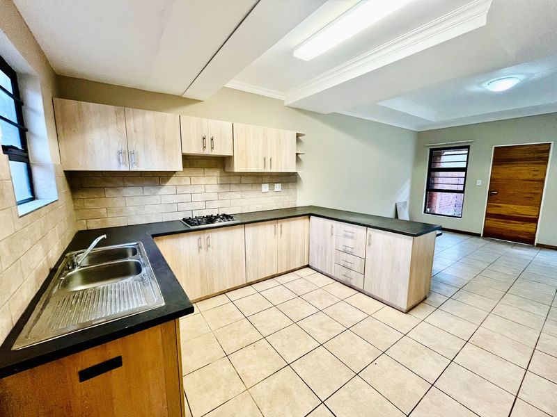 A clean, functional kitchen featuring light wood-finish cabinetry, dark countertops, a gas hob, and neutral tiled flooring in an open-plan layout.