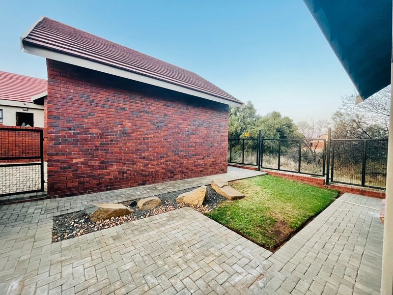 A well-maintained rear exterior area featuring a face brick townhouse wall, paved patio, small lawn, and secure metal fencing.