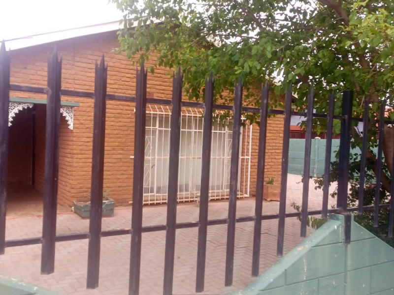 Front view of a brick house featuring a paved driveway, secure perimeter fencing, and security bars on windows.