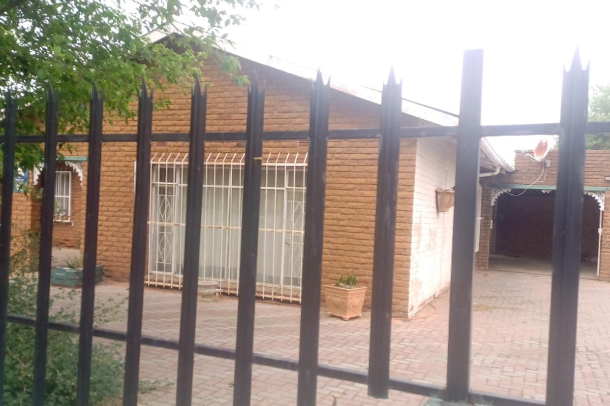 Front view of a single-story brick house featuring a paved driveway, carport, and security fencing with protective bars on windows.