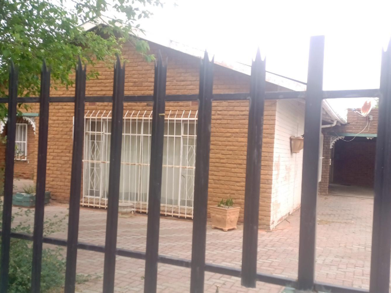 Front view of a single-story brick house featuring a paved driveway, carport, and security fencing with protective bars on windows.