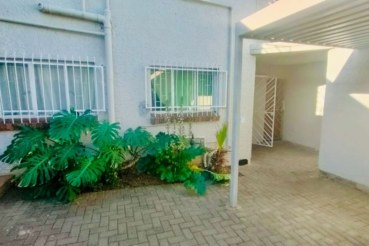 Rear exterior view of the flat featuring a paved patio area, covered walkway, and security bars on the windows.