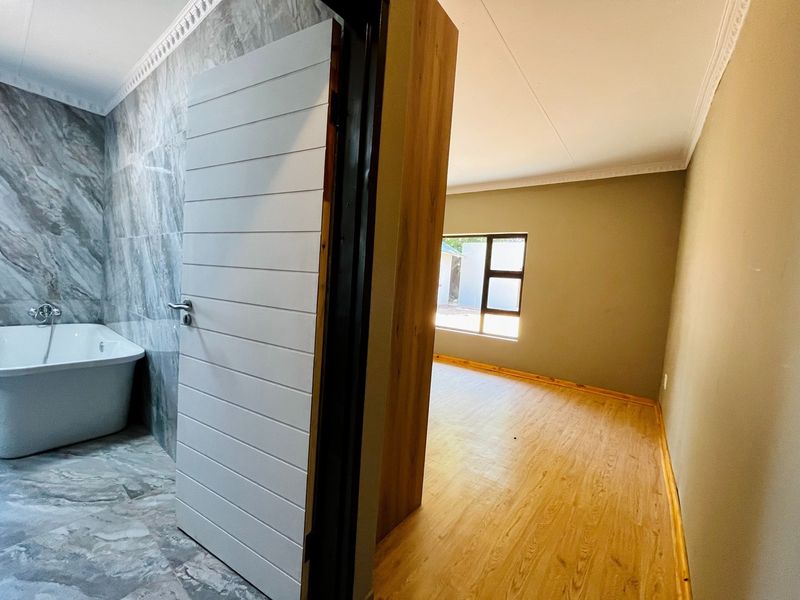 A bright, empty bedroom featuring laminate flooring and a view into an adjacent modern bathroom with marble-look tiling and a freestanding bathtub.