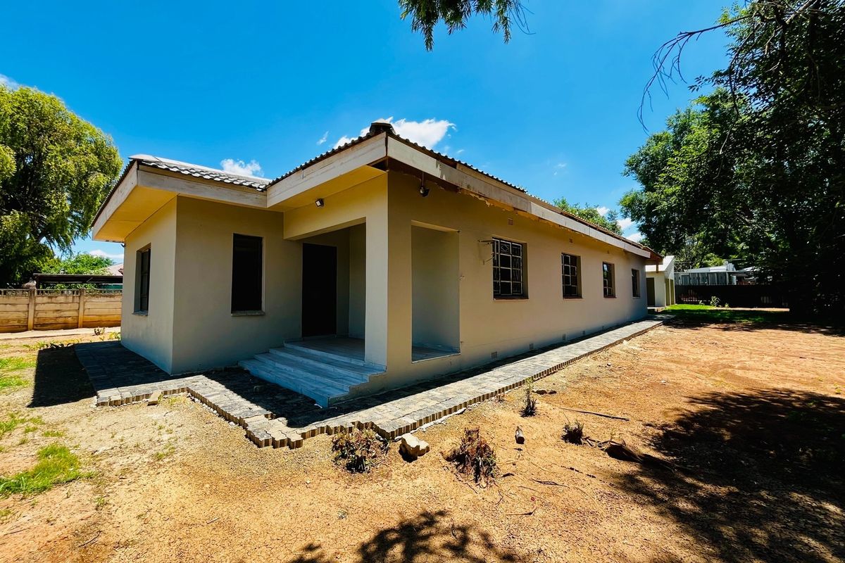 Rear view of a single-story house featuring a covered patio, tiled roof, and paved walkway, set within a large, undeveloped garden area.