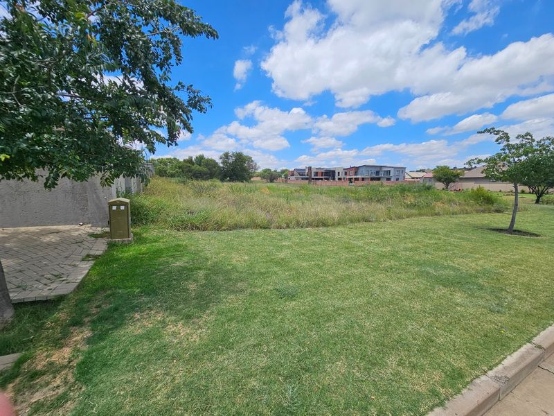A view of a vacant residential plot featuring a well-maintained lawn, paved walkway, and utility connection point in a suburban estate setting.
