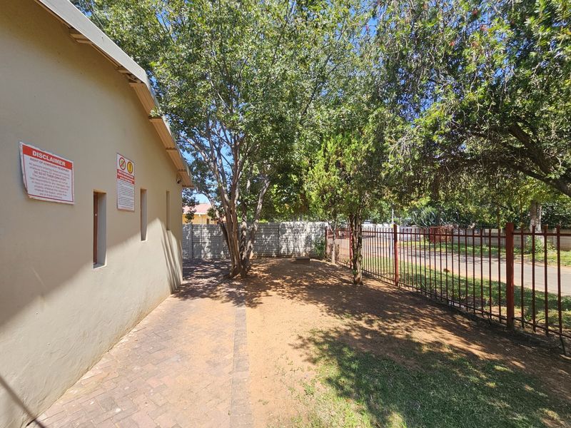 A side exterior view showing a paved walkway, mature trees, and a metal perimeter fence along the property boundary.