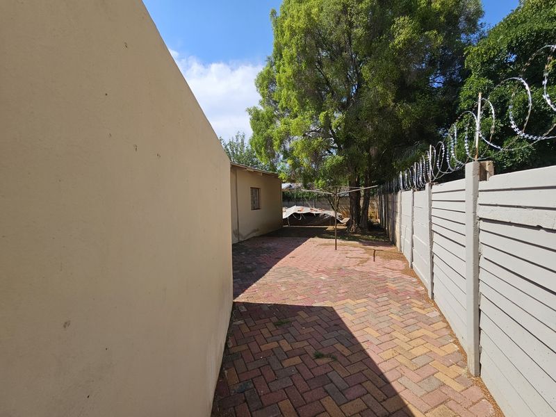 A side exterior view of the property featuring paved ground, a precast concrete boundary wall with razor wire, and mature trees.