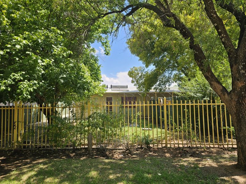 Front view of the property featuring a steel palisade fence with security razor wire, mature trees, and a roof-mounted solar water heating system.