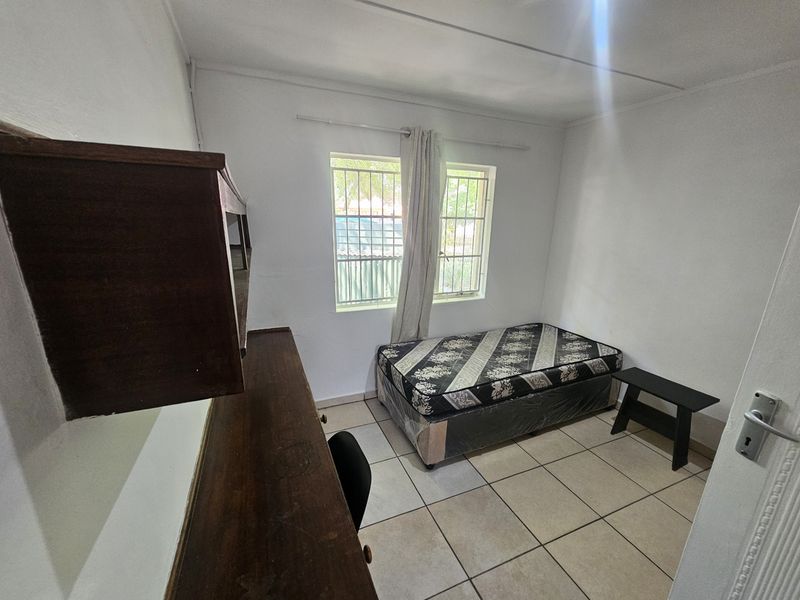 A compact bedroom featuring tiled flooring, security bars on the windows, and a built-in desk unit.