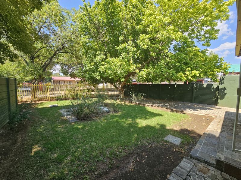 A private rear garden featuring a paved patio area, established lawn, and mature trees enclosed by perimeter fencing.