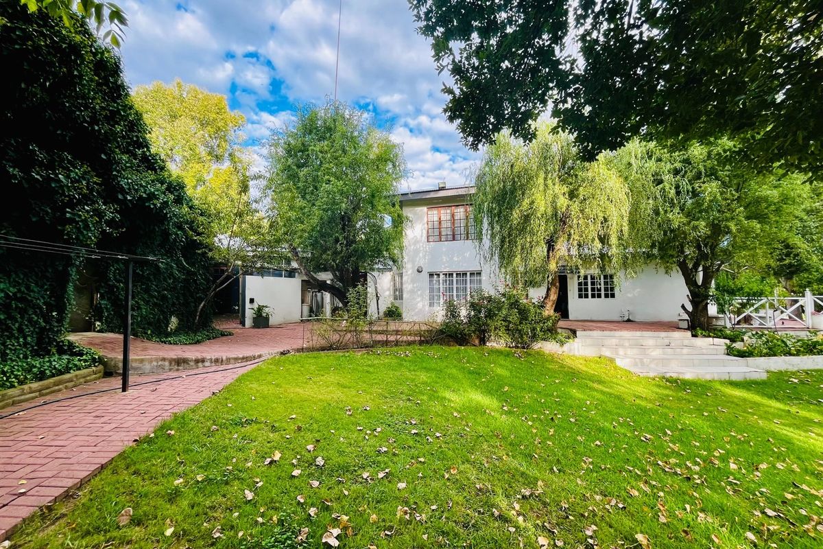A spacious rear garden featuring a lush lawn, mature trees, and brick-paved pathways leading to the white-painted rear facade of the house.