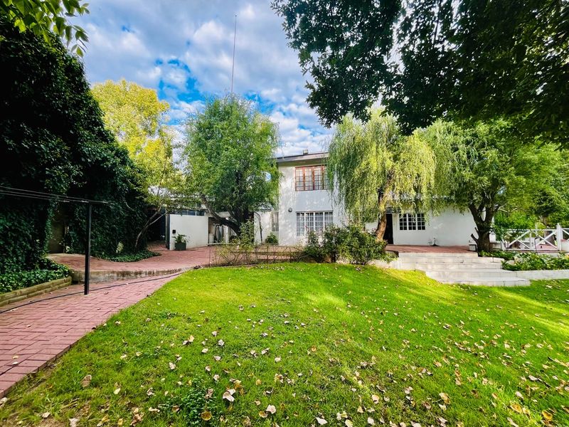 A spacious rear garden featuring a lush lawn, mature trees, and brick-paved pathways leading to the white-painted rear facade of the house.